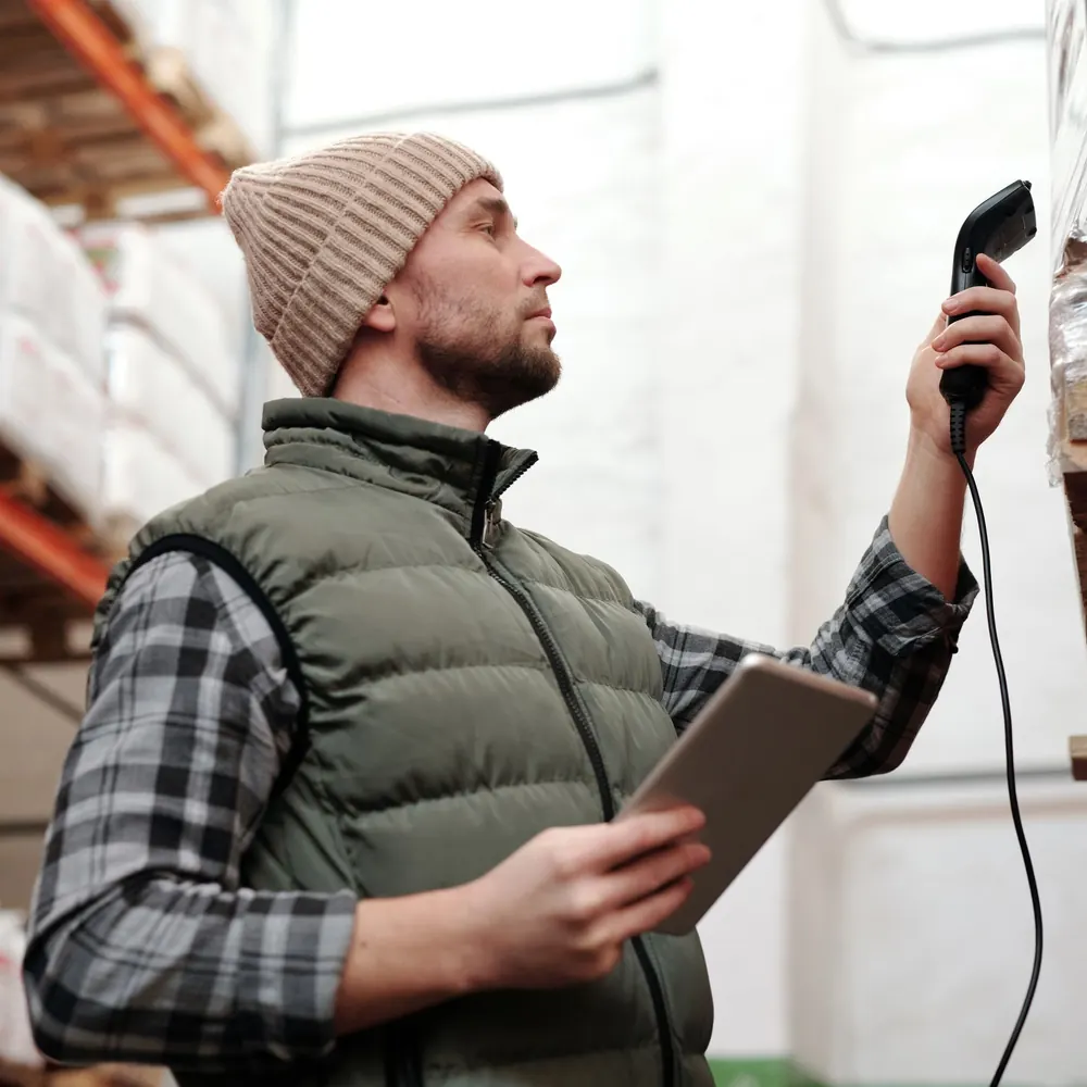 A warehouse worker in a beanie and a padded vest holds a handheld barcode scanner up to a rack label, with a tablet in his other hand.